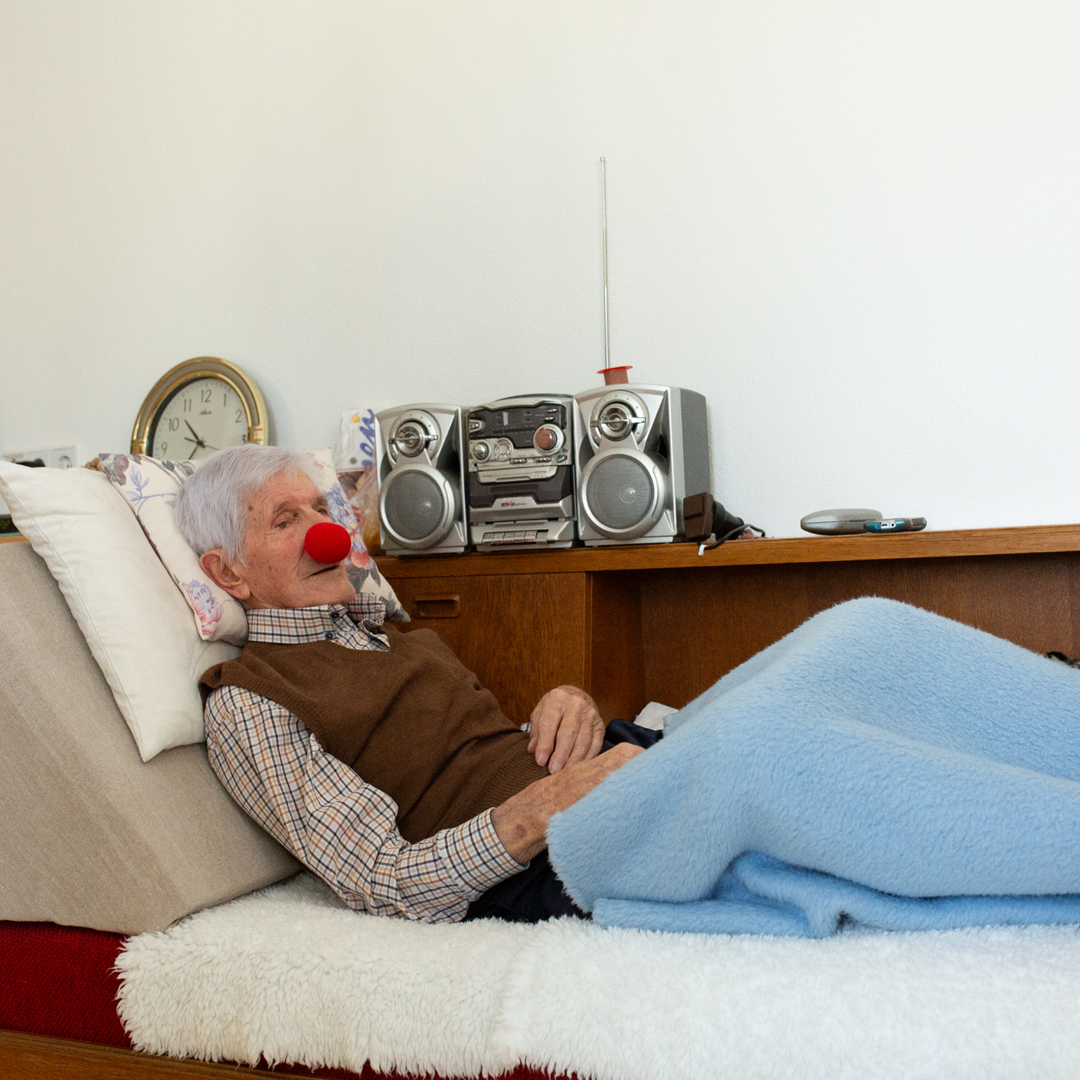 Ein älterer Herr liegt in einem Bett und trägt eine rote Schaumstoffnase.