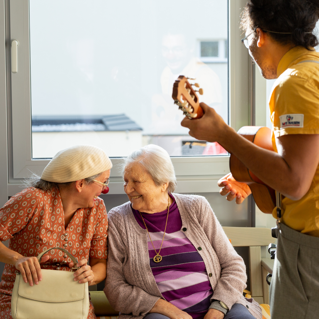 Eine Clownin unterhält sich mit einer älteren Dame, während ein Clown Ukulele spielt.