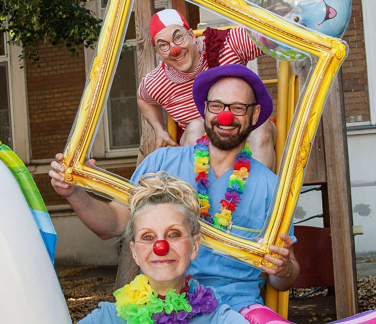 Clownin in einem Plastikeinhorn sitzt vor einem Mitarbeiter des Krankenhauses der einen goldenen Rahmen hält, dahinter steht ein weiterer Clown mit einem Plastikball 