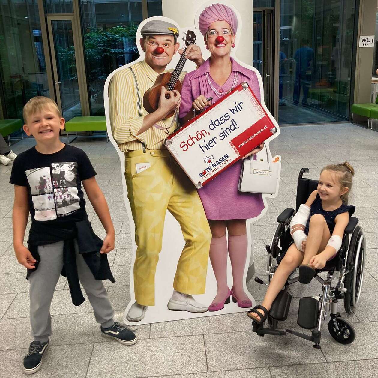 Frida und ihr Bruder im Spital vor einem Pappaufsteller mit zwei ROTE NASEN Clowns. 