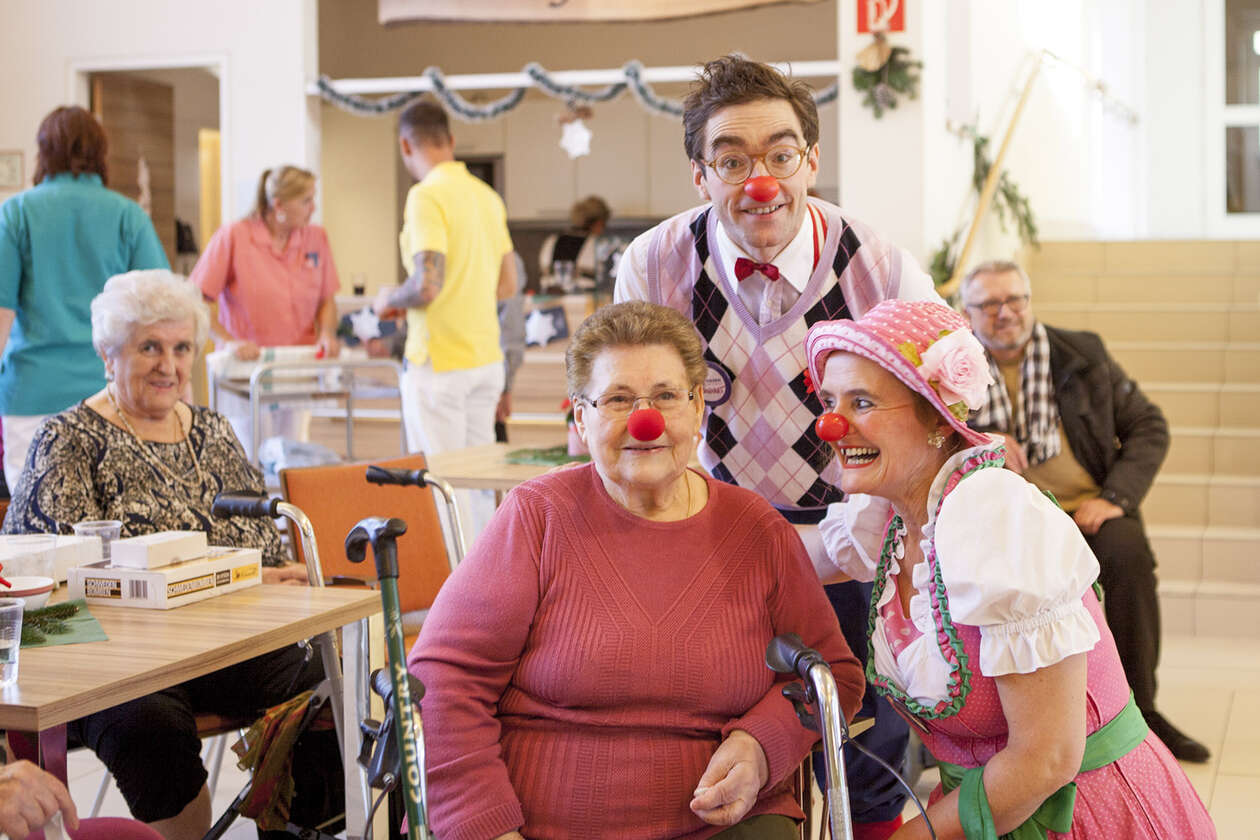 Seniorin im Rollstuhl trägt rote Schaumstoffnase, Clownin kniet sich rechts neben sie, Clown steht lachend dahinter