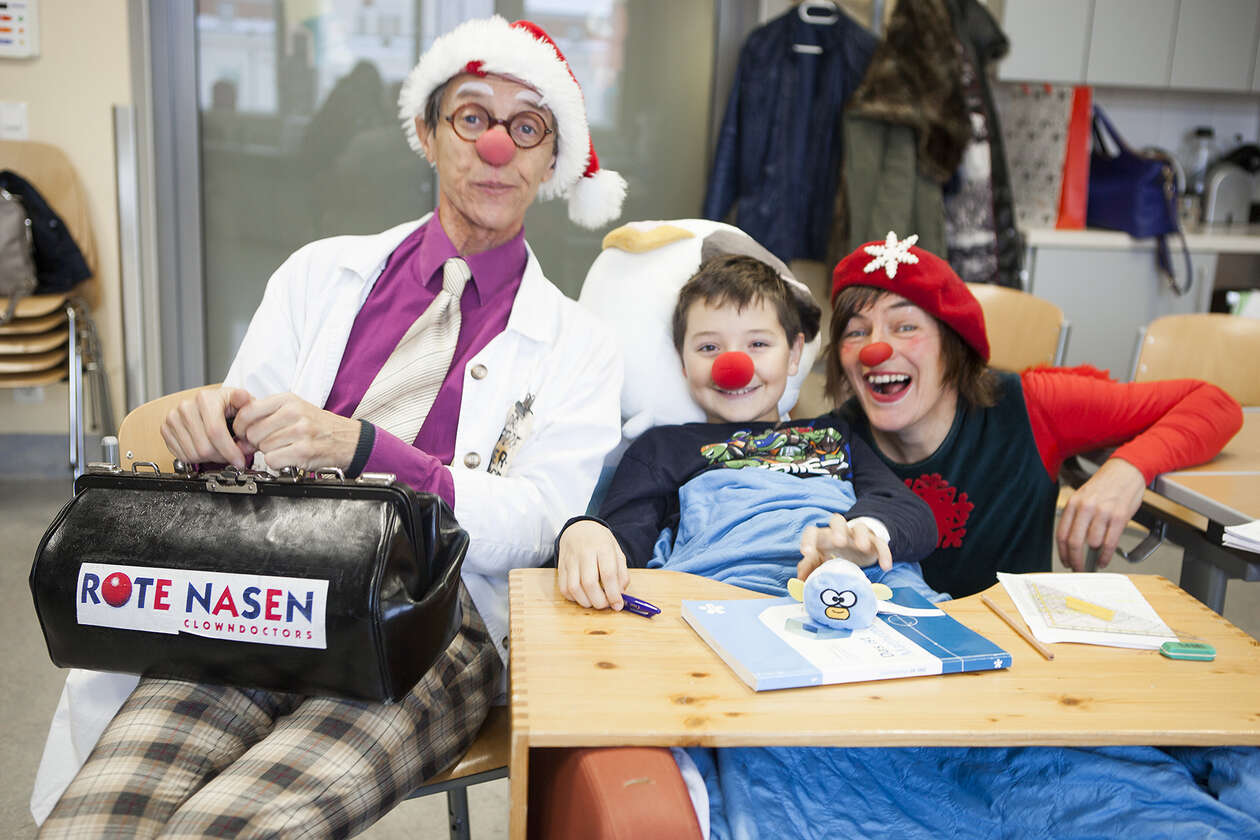 Clown mit Aktentasche am Schoß sitzt neben kleinem Jungen, Clownin sitzt rechts neben dem Jungen und lacht