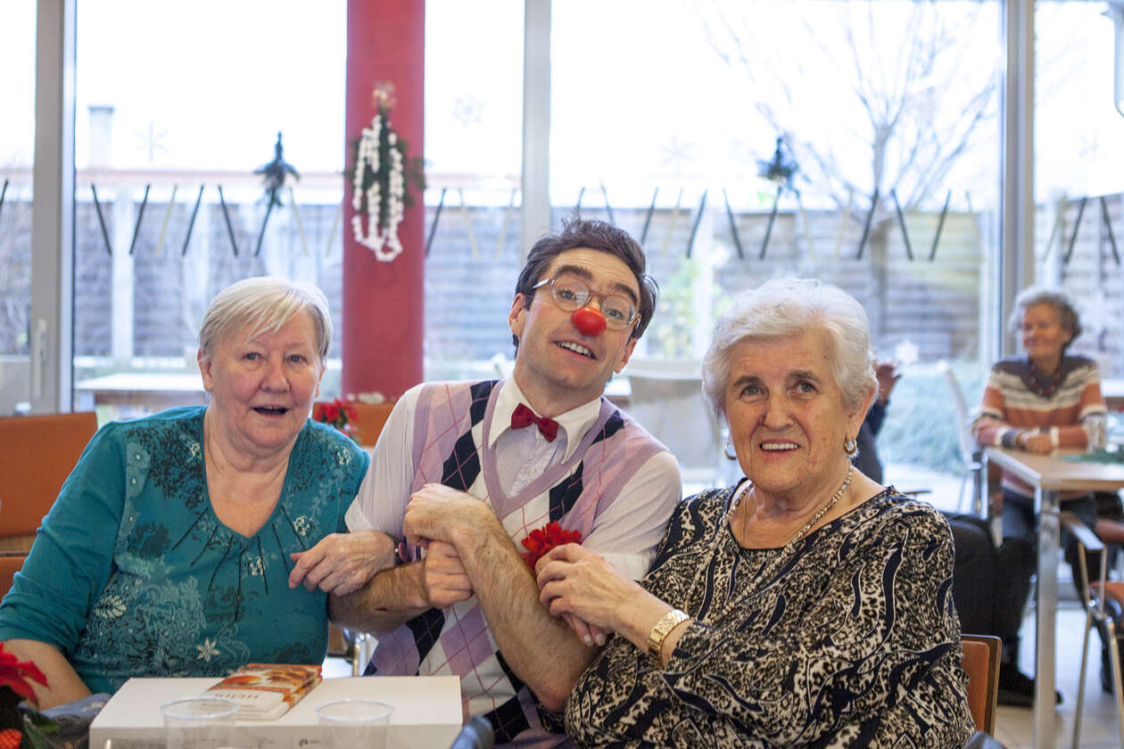 Clown sitzt zwischen zwei Seniorinnen, sie haken die Hände ein und sind dabei zu schunkeln